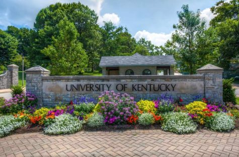 The entrance to the University of Kentuckys front campus near the Gatton School of Business and Economics and the Main Building is shown in spring. Landscaped flowering plants in purples, magenta, yellow, scarlett, orange and white are planted in front of the stone entrance marker that says University of Kentucky. The sky is blue and the sun shines bright.