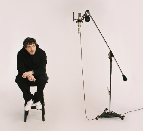 Louisville Rapper Jack Harlow in a black hoodie, black jeans, white shoes, sits on a black studio stool before a light backdrop. A high-end microphone mounted on a rolling gooseneck stand is four feet away, with a set of headphones resting on the stand and a pop filter mounted on the mic.