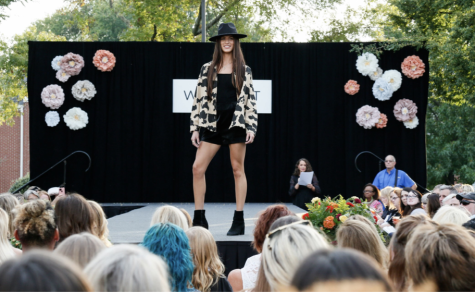 A model at a Westport Village shopping center fashion show stands onstage in a black miniskirt dress, black booties and a leopard print cardigan. Her long brunette hair is topped by a black felt brimmed hat.